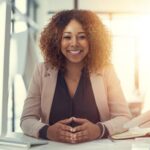 Confident professional woman smiling at her desk with calm, grounded presence in a sunlit office.