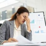 Professional woman sitting at a desk, holding her forehead in fatigue while reviewing documents, showing signs of workplace overwhelm.