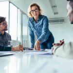 Professional woman leading a meeting, leaning over a conference table while speaking to colleagues, illustrating leadership and focus during workplace change.